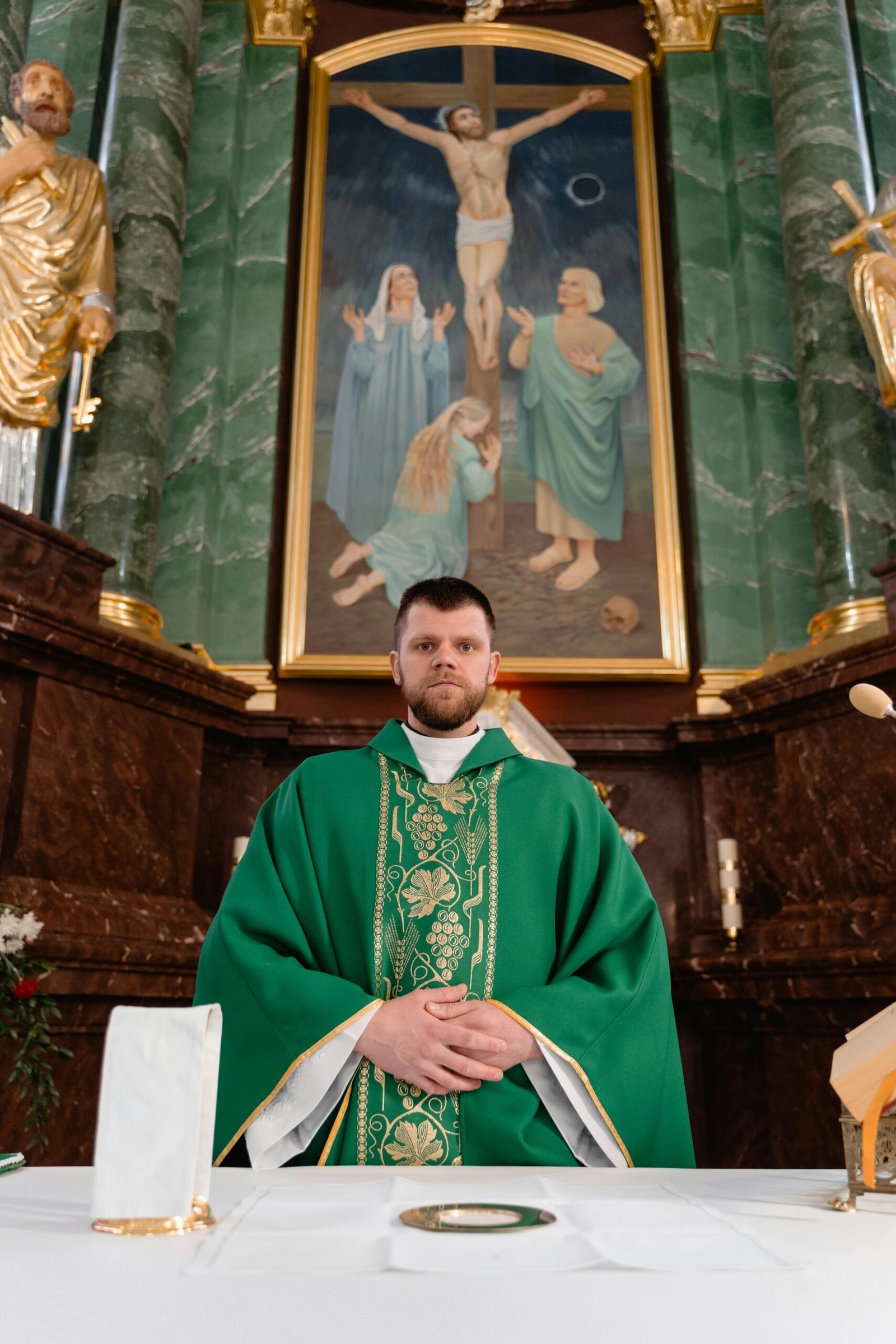 Priest in green vestment conducts religious ceremony at ornately decorated altar with crucifix.